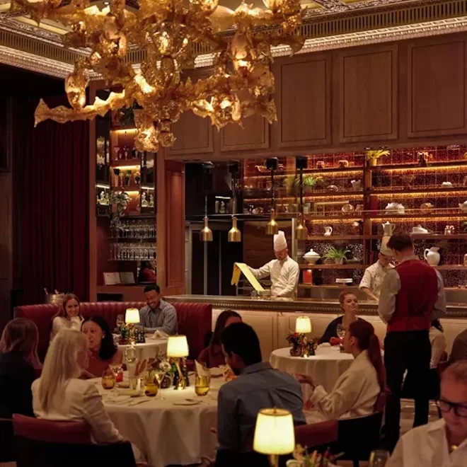 Elegant restaurant interior with several groups of people dining at round tables. Warm lighting, golden hanging decor, visible chef in the open kitchen, and a server attending to guests create a sophisticated, lively atmosphere.