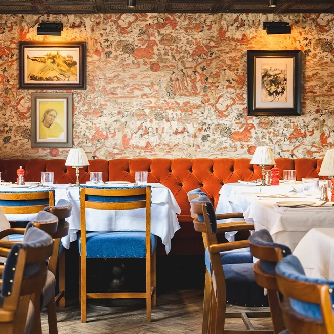 A cozy restaurant interior with white-clothed tables, wooden chairs with blue cushions, and a long orange tufted bench. The wall is decorated with patterned wallpaper and framed art. Table lamps add a warm ambiance.