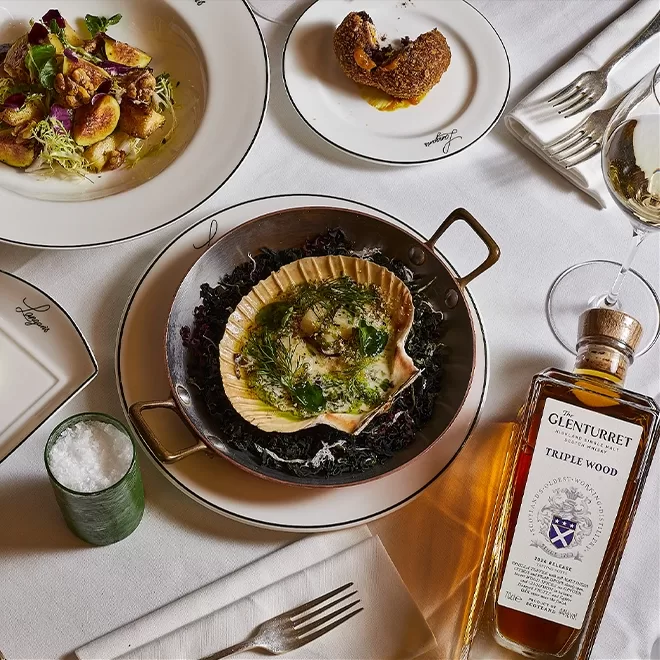 A plated scallop dish garnished with herbs, a bottle of Glenturret Triple Wood whisky, a salt bowl, cutlery, and two other elegant plates with gourmet food arranged on a white tablecloth.