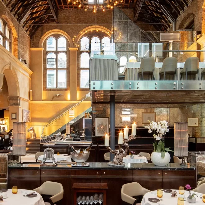 A spacious, elegant restaurant with high vaulted ceilings, large arched windows, a mezzanine level, and stylish decor including orchids, candles, and neatly set tables. Warm lighting creates a welcoming atmosphere.