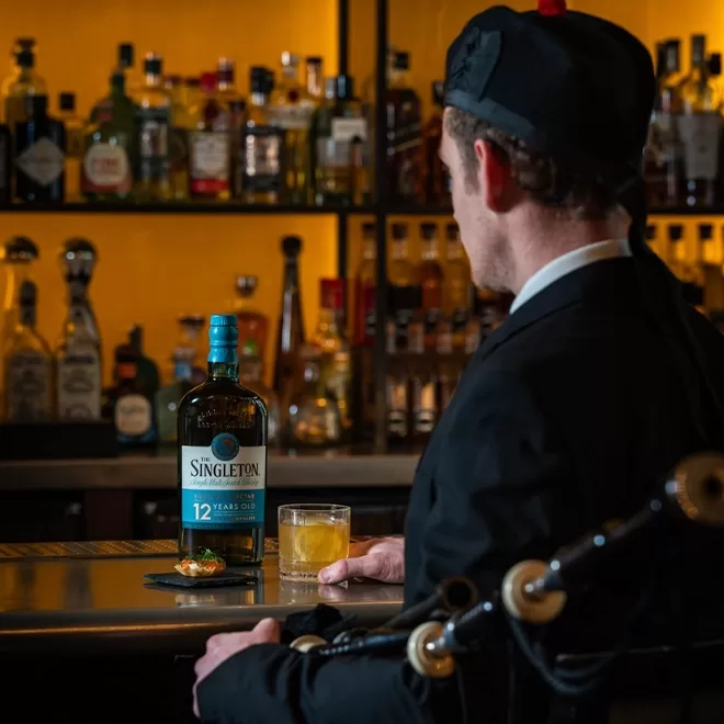 A man in traditional attire holding bagpipes sits at a bar, facing a bottle of The Singleton 12-year-old whisky and a glass of whisky, with shelves of liquor bottles in the background.