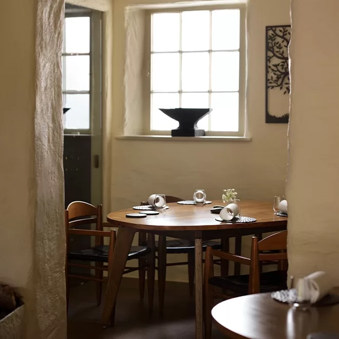 A cozy dining room with a round wooden table set for four, surrounded by wooden chairs. Soft natural light streams through a window, revealing minimalist decor and a black vase on the windowsill.