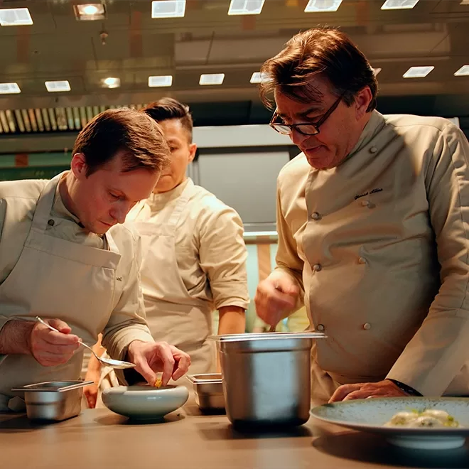 Three chefs in beige uniforms work together in a kitchen, carefully plating food. One chef focuses on adding a finishing touch to a dish while the others observe and assist. Stainless steel pots and bowls are on the counter.