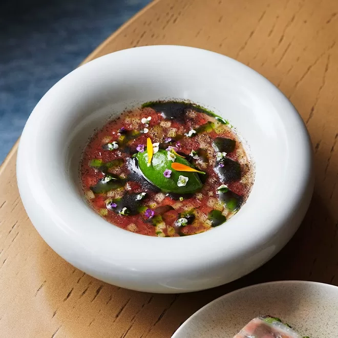 A white bowl with a colorful, artistic dish featuring a green mousse or puree in the center, surrounded by a light broth, diced vegetables, edible flowers, and drops of dark and green sauce, on a wooden table.