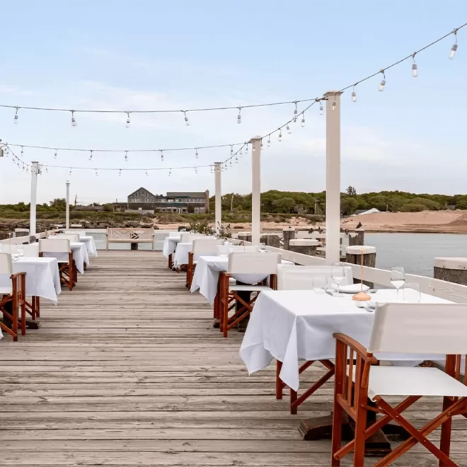 Outdoor restaurant on a wooden dock with white tablecloth-covered tables and wooden chairs, string lights overhead, overlooking water and greenery under a clear sky.