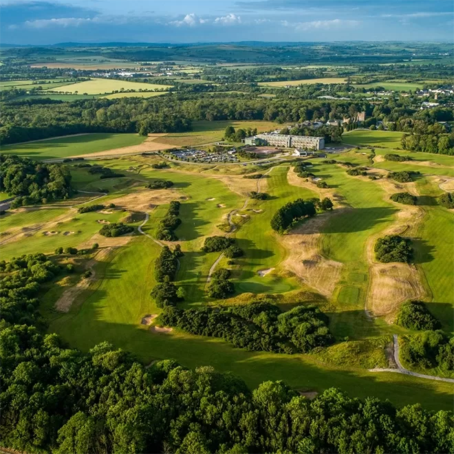 Aerial view of a golf course surrounded by green fields, trees, and a few buildings, with hills and farmland stretching into the distance under a partly cloudy sky.