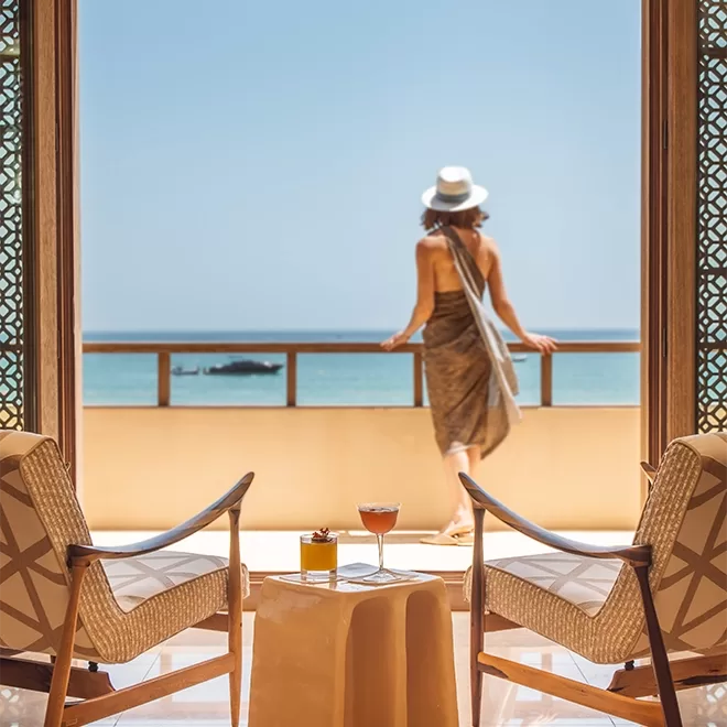 A woman in a sun hat and dress stands on a balcony overlooking the ocean. Two chairs and a small table with a drink and book are in the foreground, framed by ornate wooden doors. The sky is clear and blue in Temuda Bay.