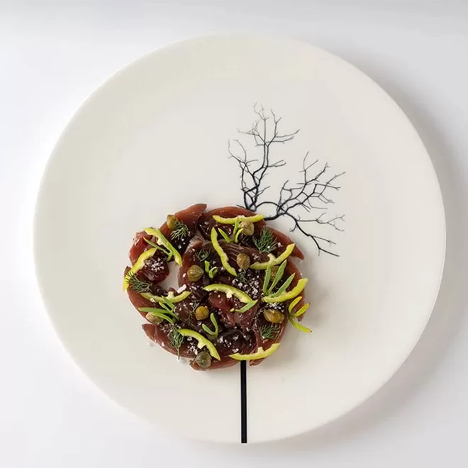 A round dish of artfully arranged food sits on a white plate, resembling the foliage of a tree, with a black tree trunk and branches design printed on the plate extending above it.