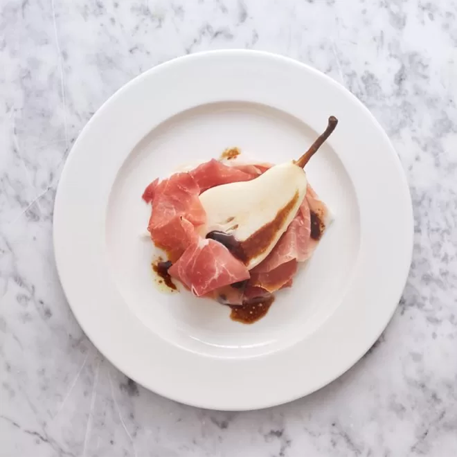 A white plate on a marble surface holds slices of prosciutto wrapped around a halved pear, drizzled with a dark balsamic glaze.