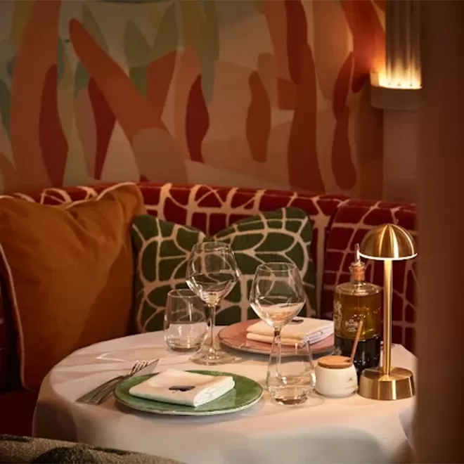 A cozy restaurant table set for two, with plates, glasses, and napkins on a white tablecloth, surrounded by patterned cushions and lit by a small golden lamp, with abstract wall art in the background.