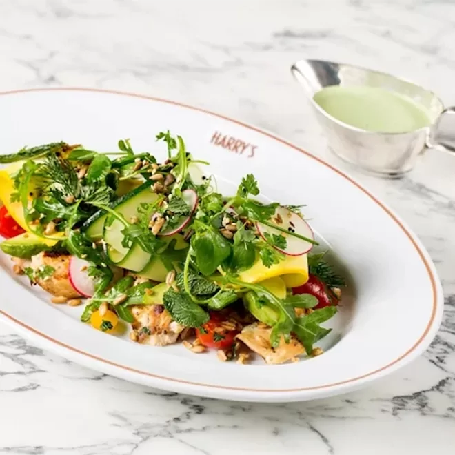 A plate of fresh salad with grilled chicken, avocado, mango slices, radishes, greens, and seeds, served on a white dish labeled HARRYS, with a side of green dressing in a small pitcher on a marble table.