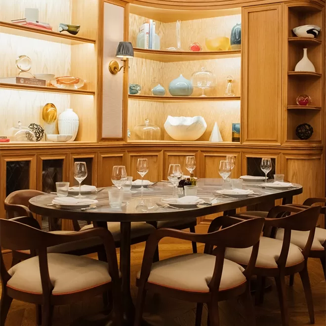 A modern dining area with a marble table set for six, surrounded by wooden chairs with cream cushions. Behind, a wooden display shelf holds decorative vases, bowls, and books, softly lit for a cozy ambiance.