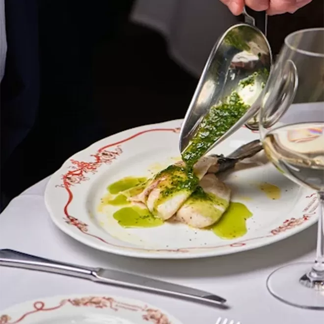 A person pours green herb sauce from a metal ladle over a plated fish fillet on a decorative white plate, set on a white tablecloth next to a glass of white wine, fork, and knife.