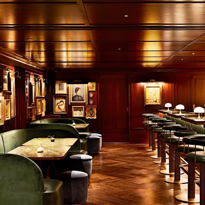 A sophisticated bar with dark wood paneling, green velvet booths, marble tables, and high stools; vintage portraits and artwork decorate the walls, creating an elegant, intimate atmosphere.