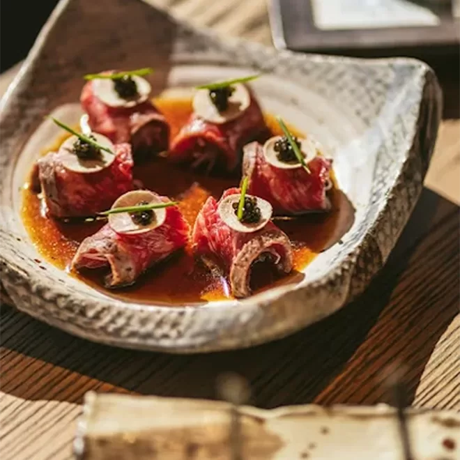 Five pieces of thinly sliced beef wrapped around a filling, topped with a round garnish and green herb, served on a ceramic plate with a brown sauce.