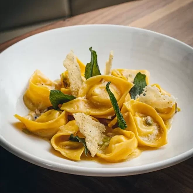 A white bowl filled with tortellini pasta topped with crispy sage leaves and thin Parmesan crisps, set on a wooden table.