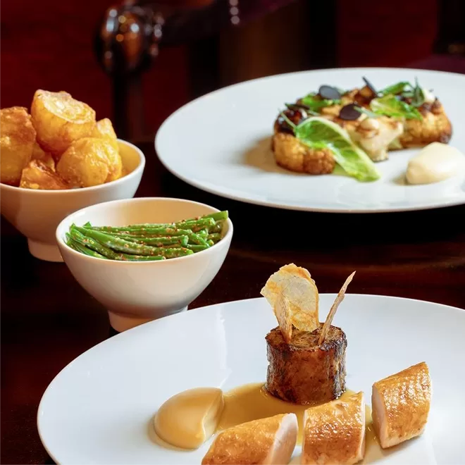 A gourmet meal is artfully plated on white dishes, featuring roast meat, pureed sides, crispy potatoes, green beans, and a vegetable-based dish garnished with fresh greens and sauce. The setting is elegant and refined.
