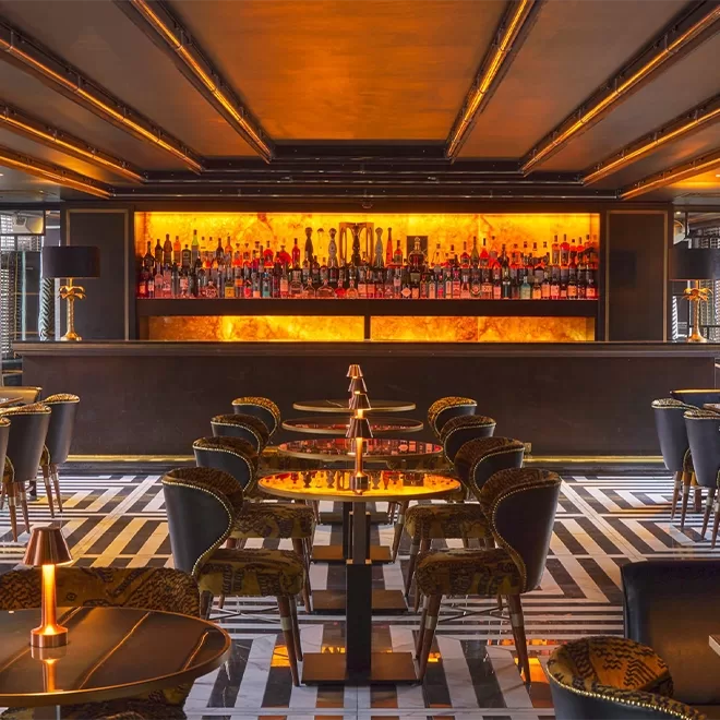 A stylish modern bar with patterned flooring, dark chairs, and glowing orange lighting illuminating shelves of bottles behind a sleek black bar counter. Small lamps are on each table, creating a warm, inviting atmosphere.