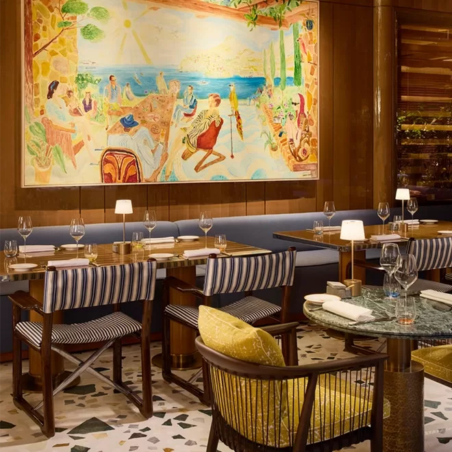 A stylish restaurant interior with striped chairs, a marble table, set tables with wine glasses, and a large colorful painting of a lively beach scene hanging on a wooden-paneled wall.