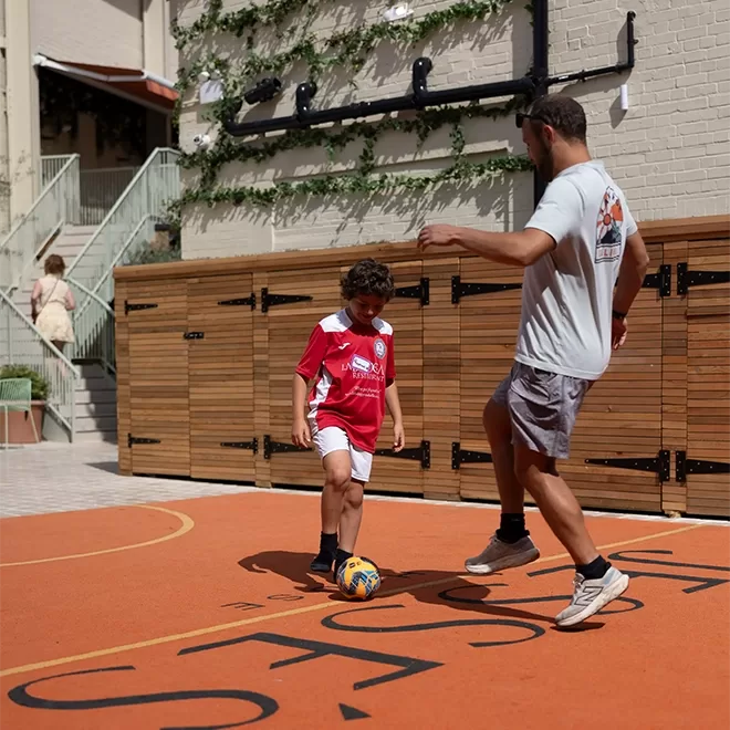 A young boy in a red soccer jersey and an adult in a gray shirt play soccer together on an orange outdoor court, with wooden lockers and a flight of stairs visible in the background.