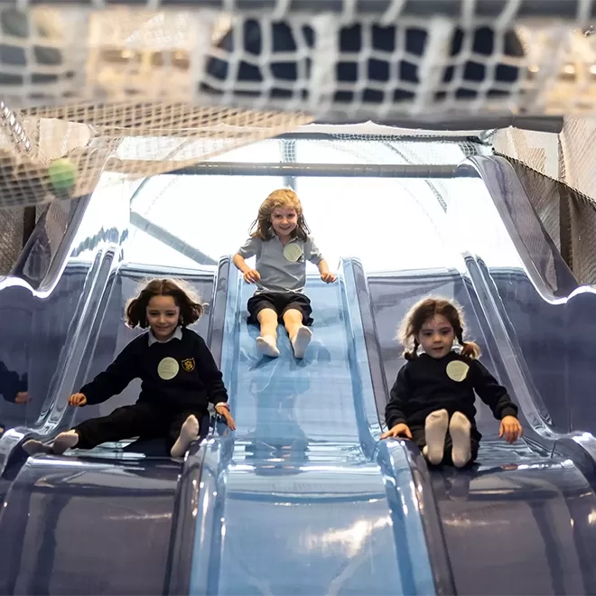 Three young children in school uniforms slide down adjacent blue slides indoors, smiling and having fun. A safety net is visible overhead in the play area.