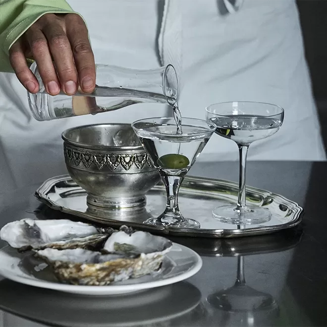 A person pours a clear liquid from a glass carafe into a cocktail glass with an olive on a silver tray, next to another glass, a metal bowl, and a plate with open oysters.