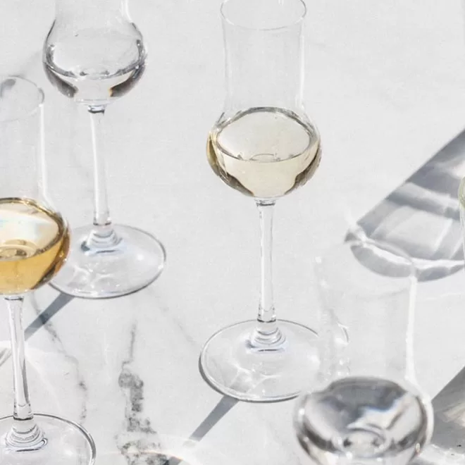 Several clear, stemmed glasses, some filled with light golden and transparent liquid, are arranged on a white marble surface with sunlight casting shadows and reflections across the scene.