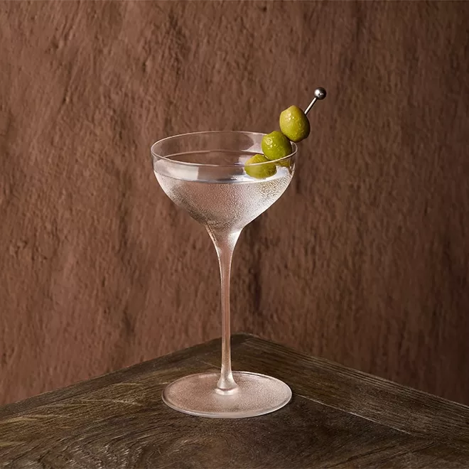 A martini glass filled with a clear cocktail, garnished with three green olives on a metal pick, sits on a dark wooden surface against a textured brown background.
