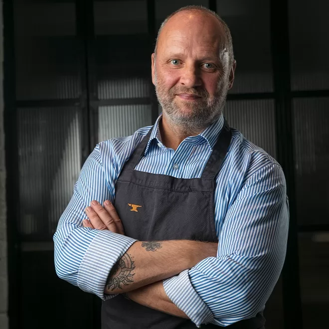 Chef Simon Rogan stands confidently with arms crossed, wearing a striped shirt and dark apron. A tattoo is visible on his forearm. The background is dark with vertical lines.