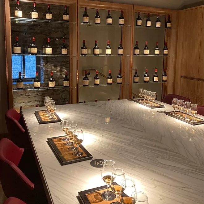 A modern tasting room with a large white marble table set with wine glasses and snacks, surrounded by shelves displaying multiple bottles of wine or liquor.