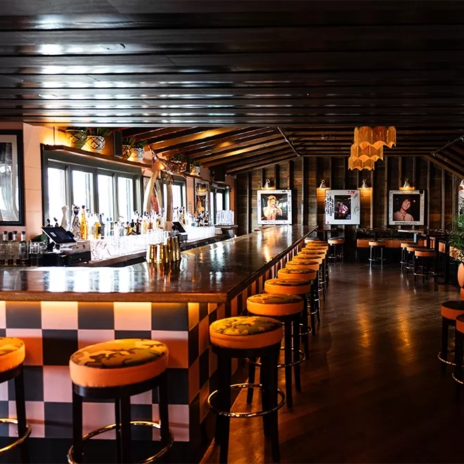 A stylish bar with a long counter, cushioned stools, wooden floors, and exposed beam ceiling. Large windows line one wall, and framed photos and pendant lights add to the cozy, upscale atmosphere.