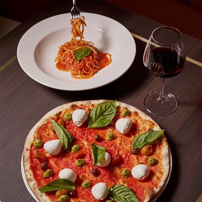 A table with a Margherita pizza topped with mozzarella, basil, and pesto, a plate of spaghetti with tomato sauce being twirled on a fork, and a glass of red wine.