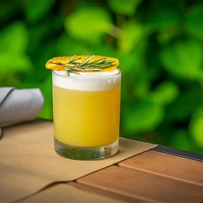 A yellow cocktail with a frothy top served in a short glass, garnished with a dried orange slice and rosemary sprig, sits on a table with a beige placemat and green foliage in the background.