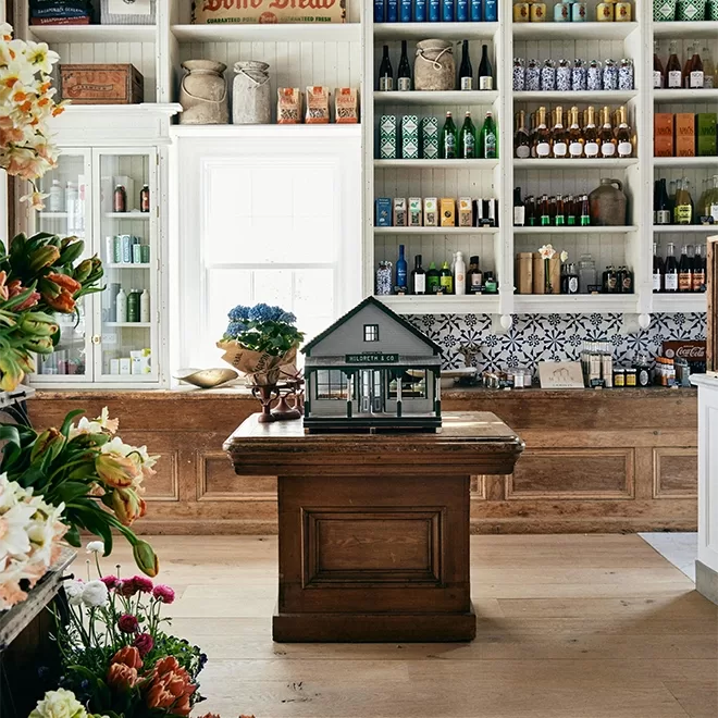 A cozy shop interior with shelves full of bottles, jars, and goods; a wooden counter runs along the back, and a small model house sits on a central table. Fresh flowers decorate the scene.