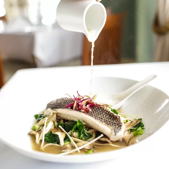 A close-up of a white bowl with steamed fish fillet, leafy greens, and mushrooms, as hot broth is being poured over the dish from a small white pitcher. Steam rises from the food.