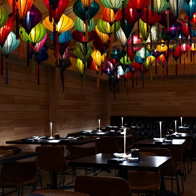 A dimly lit restaurant with wooden walls and dark tables. Colorful, lantern-style lights hang from the ceiling, creating a vibrant and cozy atmosphere over the dining area.