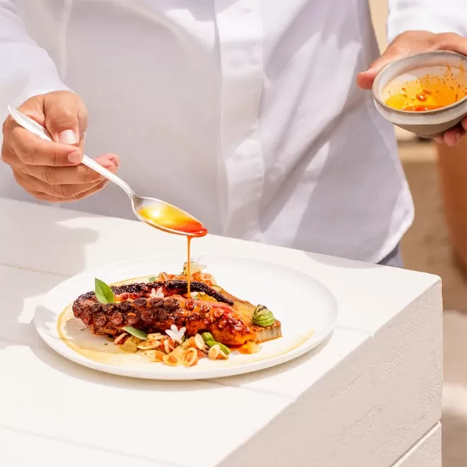 A person in a white shirt drizzles sauce from a spoon onto a plated dish of grilled meat garnished with herbs and vegetables, outdoors on a sunny day.