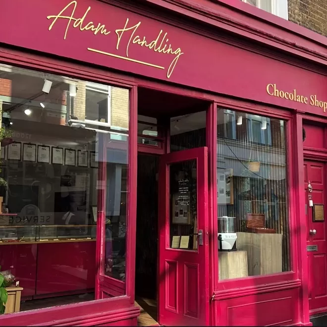 A vibrant red storefront with gold lettering reading Adam Handling and Chocolate Shop above a glass door and windows, revealing a glimpse of the interior.