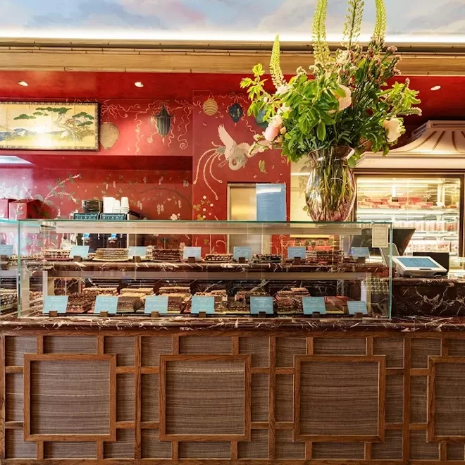 A bakery counter with assorted pastries displayed behind glass, a vase of tall colorful flowers on top, and a red wall with bird and floral art in the background. The ceiling is painted to resemble the sky.