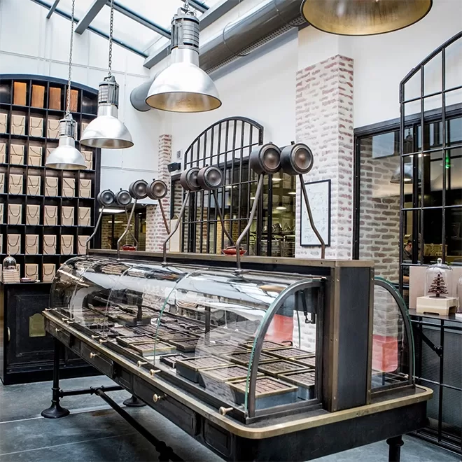 A modern, industrial-style bakery or chocolate shop with a large glass display case holding assorted treats under metal pendant lights, exposed brick walls, large windows, and shelves with paper packets.