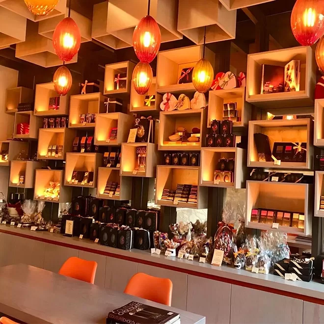 A display wall of shelves filled with assorted chocolates and gift boxes in a modern chocolate shop, warmly lit by hanging orange lamps above a counter with more treats and orange chairs.