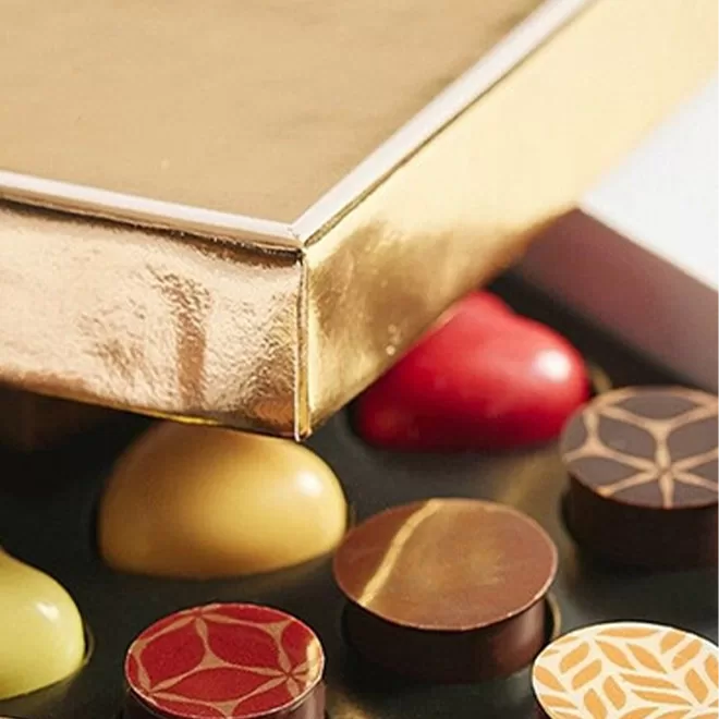 A close-up of assorted gourmet chocolates with colorful designs next to a shiny gold-edged box.