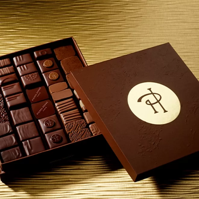 An open box of assorted chocolates in a brown, textured box with a gold circular logo on the lid, placed on a gold-striped surface.
