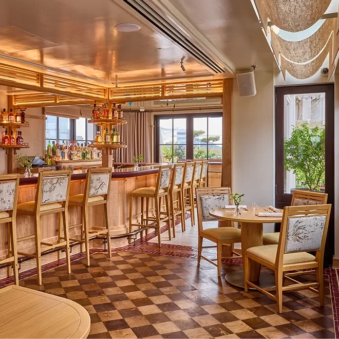 A stylish bar with wooden furniture, patterned high stools, a checkered floor, and a well-stocked bar. Large windows let in natural light, and a small round dining table with chairs sits nearby.