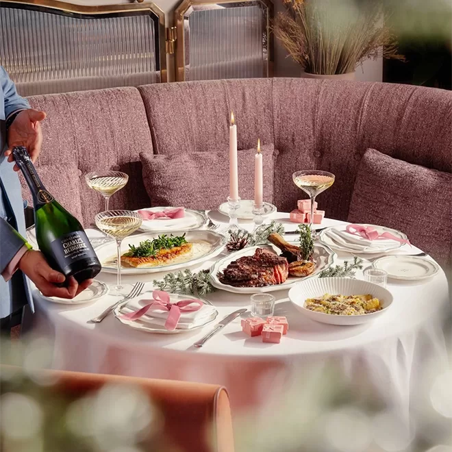 A round table set for a romantic dinner with steak, pasta, and greens, decorated with pink ribbons, candles, champagne, and gift boxes. A person is opening a champagne bottle. Plush seating and elegant tableware complete the scene.