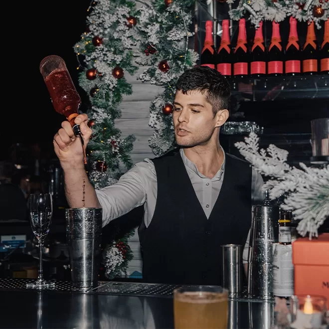 A bartender in a vest pours a red drink into a shaker at a festive bar decorated with white and red Christmas ornaments and garlands. Glasses and bottles are visible on the counter.