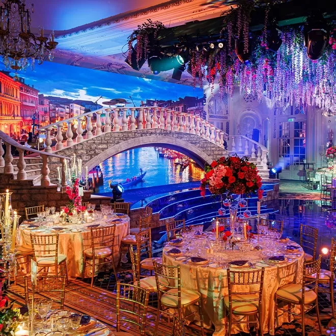 Lavish indoor event setup with round, elegant tables, gold chairs, and crystal chandeliers. The decor features a large faux canal, ornate bridge, and hanging flowers, creating a Venetian-inspired atmosphere.
