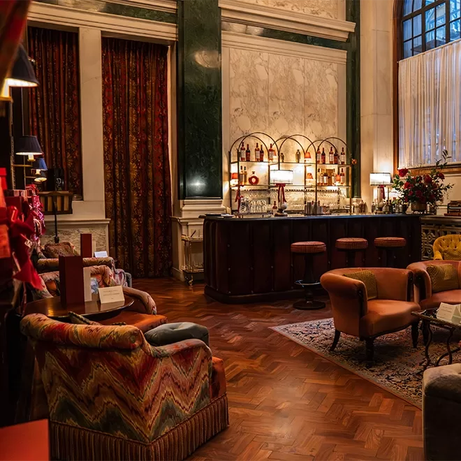 Luxurious hotel lounge with plush, colorful armchairs, a bar with bottles displayed on shelves, elegant lamps, patterned curtains, and warm lighting, featuring a large arched window and wood parquet flooring.