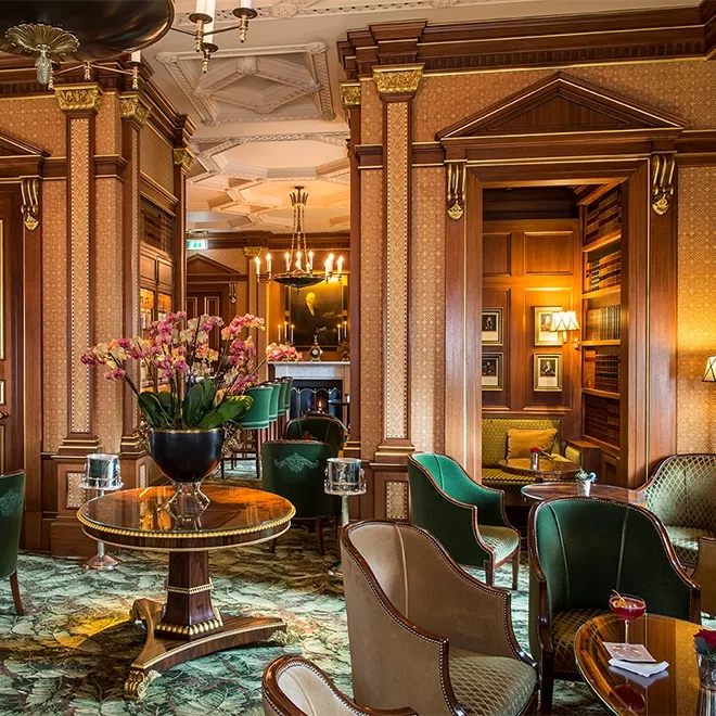 Elegant hotel lounge with green velvet chairs, wood-paneled walls, gold accents, a round table with a large flower arrangement, patterned carpet, and a cozy bookshelf nook with warm lighting.