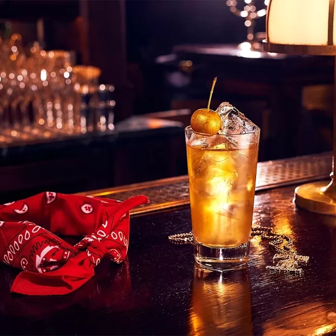 A highball glass filled with an amber cocktail, ice cubes, and garnished with a cherry, sits on a dimly lit bar beside a red bandana and a lamp. Glassware and bottles are visible in the blurred background.